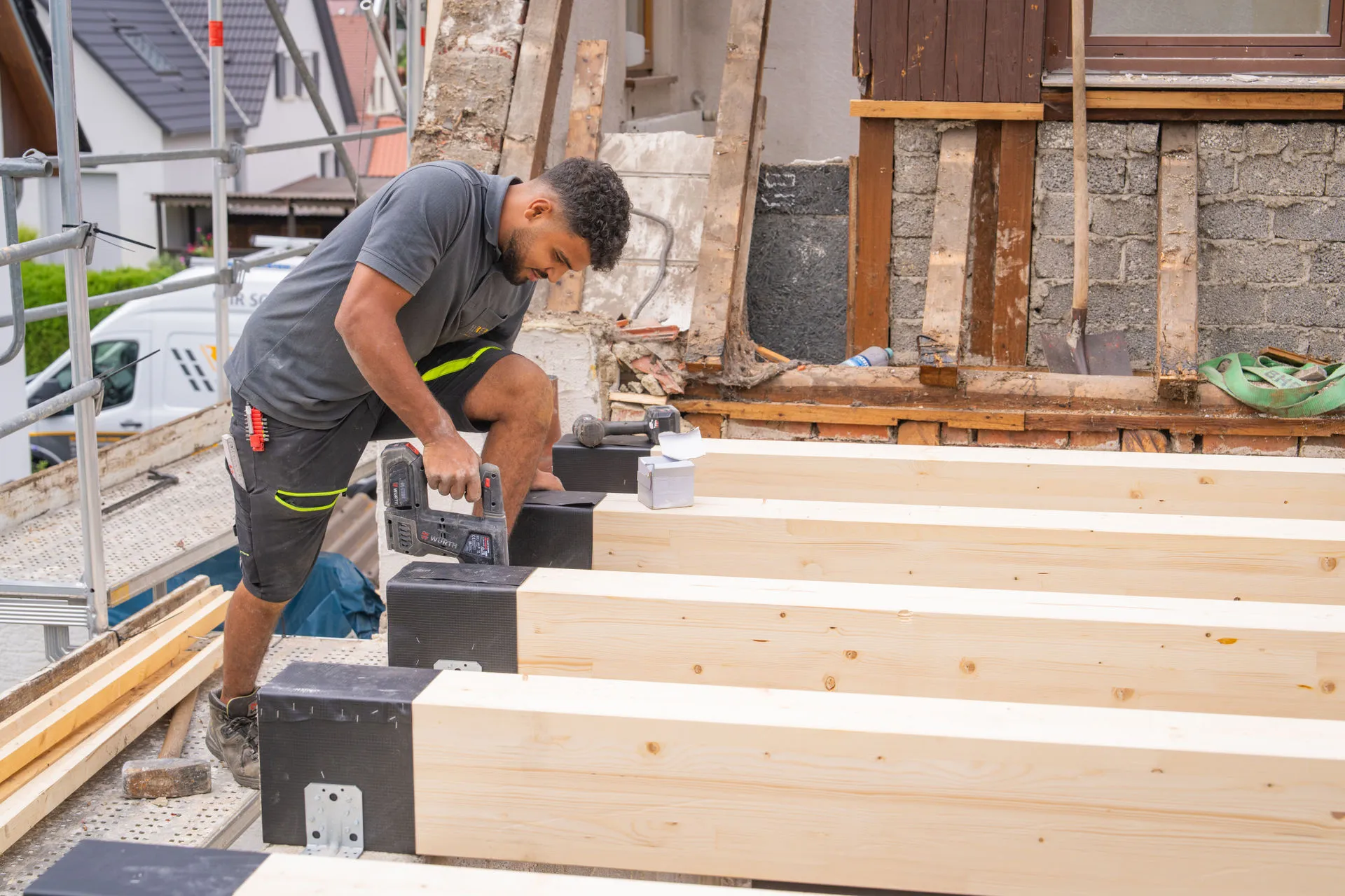 Ein Handwerker auf einer Baustelle befestigt mit einem Tacker schwarze Schutzfolie auf massiven Holzbalken einer neuen Deckenkonstruktion; im Hintergrund ist ein teils entkerntes Gebäude und ein Gerüst zu sehen.