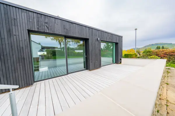 Außenansicht eines modernen Anbaus mit dunkler Holzfassade, großen Glasschiebetüren und einer hellen, großzügigen Terrasse unter bewölktem Himmel.