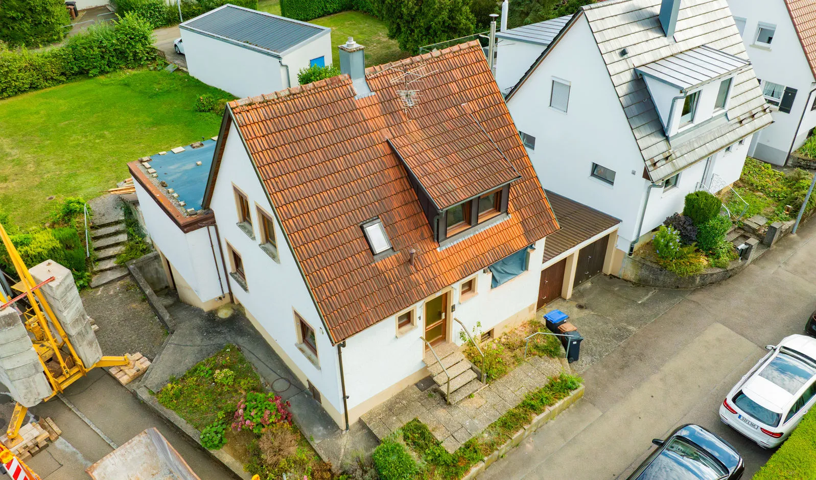 Schräge Luftaufnahme eines weißen Einfamilienhauses mit rotem Ziegeldach während einer Sanierungsphase; links im Bild ist der Fuß eines gelben Baukrans auf der Baustelle zu sehen.