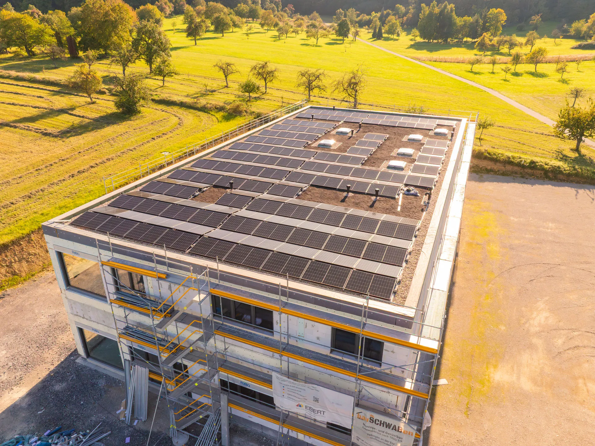Luftaufnahme eines im Bau befindlichen Gewerbegebäudes mit einer großflächigen Photovoltaikanlage auf dem Flachdach; das Gebäude ist eingerüstet und liegt in einer grünen Landschaft.