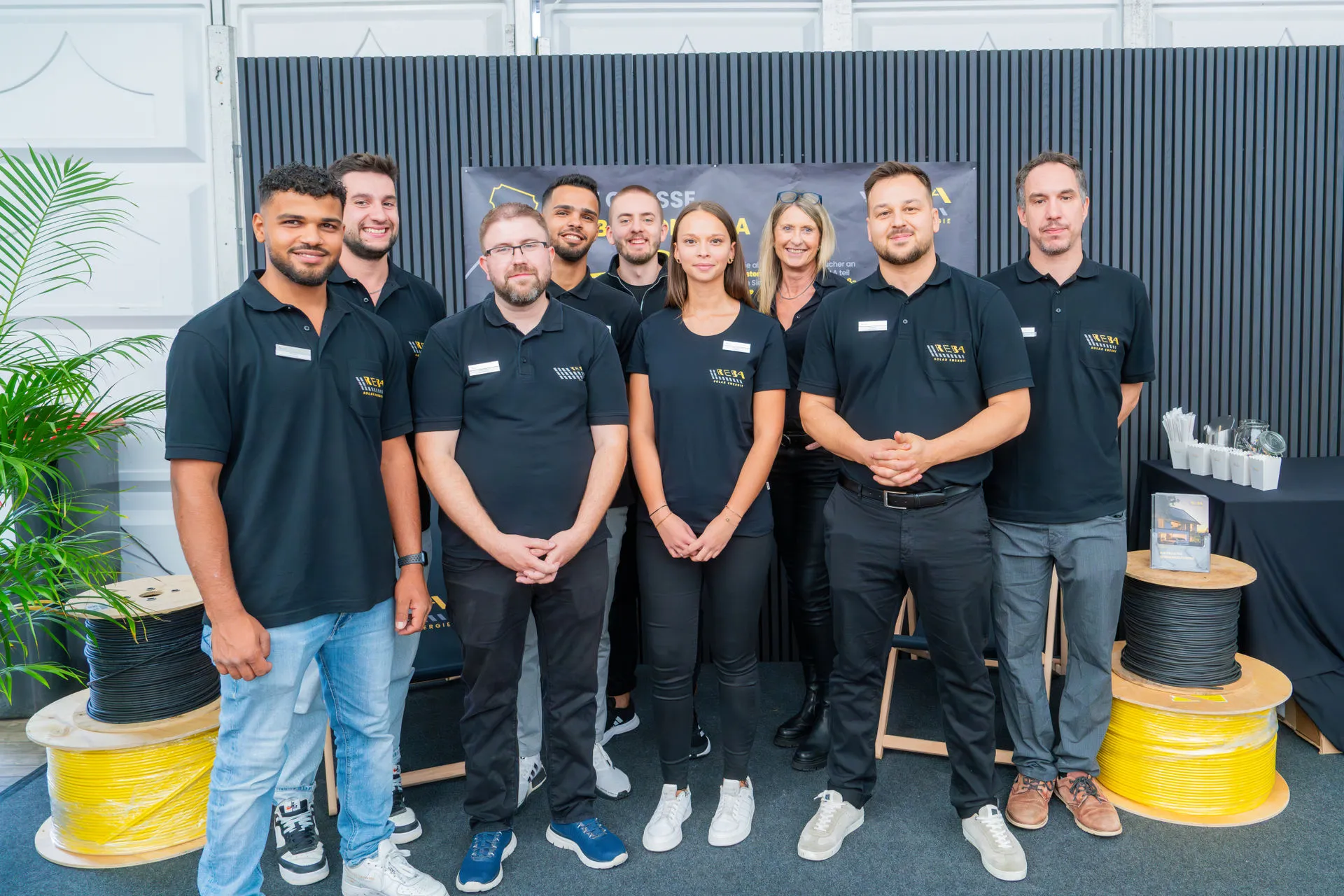 Das Team der REBA Solar Energie: Neun Mitarbeiterinnen und Mitarbeiter posieren lächelnd in einheitlicher schwarzer Firmenbekleidung mit REBA-Logo vor einem Messestand. Im Vordergrund stehen Kabeltrommeln mit gelben und schwarzen Erdkabeln.