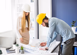 Man in a yellow hard hat and woman in a white hard hat reviewing architectural blueprints on a table in a modern office.