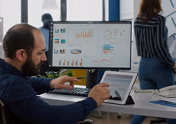 Man using a tablet and computer with data charts on screens in an office setting.