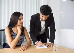 A man in a suit is writing on a notepad while a woman in a sleeveless dress watches attentively at a desk.