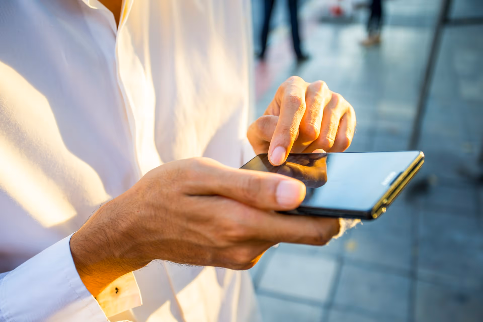 Person in white shirt using a black smartphone outdoors on a paved surface.