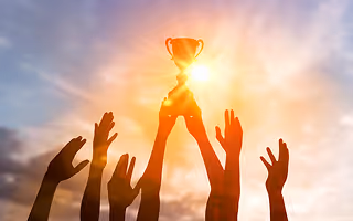 Hands reaching up together holding a glowing trophy against a bright sunset sky.