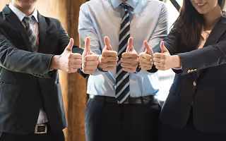 Three professionals in suits giving thumbs up with smiling expressions.