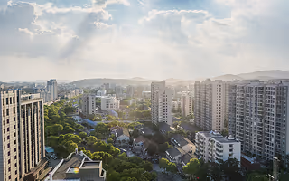 Cityscape with high-rise buildings, residential houses, and greenery under a sky with scattered clouds and sunlight rays.