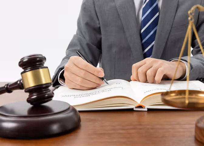 Person in suit writing in a book at a desk with a gavel and scales of justice nearby.