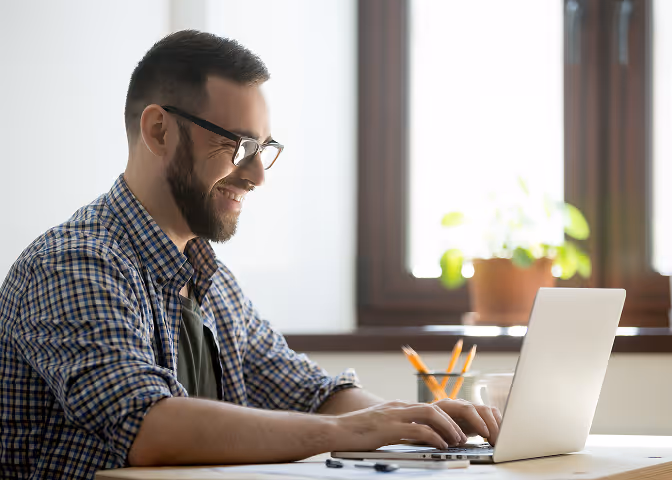 Man with beard and glasses smiling while typing on a laptop in a bright room near a window.