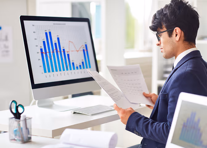Man in suit examining financial documents while viewing bar and line graphs on a desktop computer screen.
