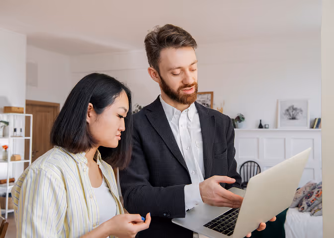 Man in suit showing a laptop screen to a woman in a striped shirt inside a modern living room.