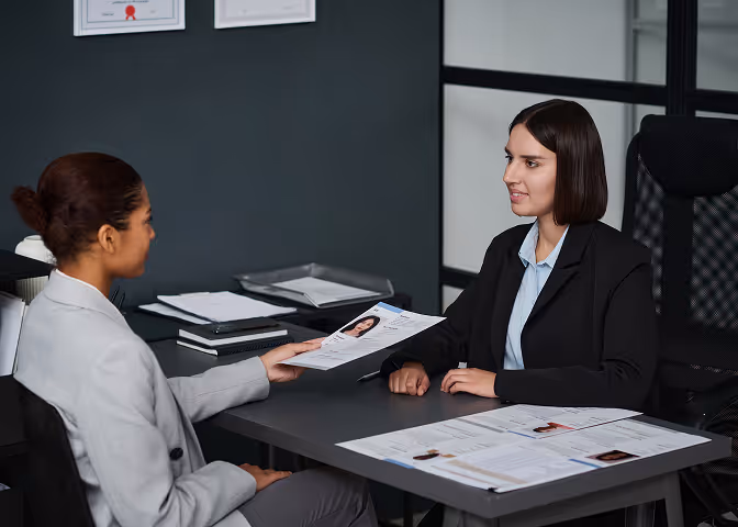 Two professional women at a desk reviewing printed resumes during a job interview.