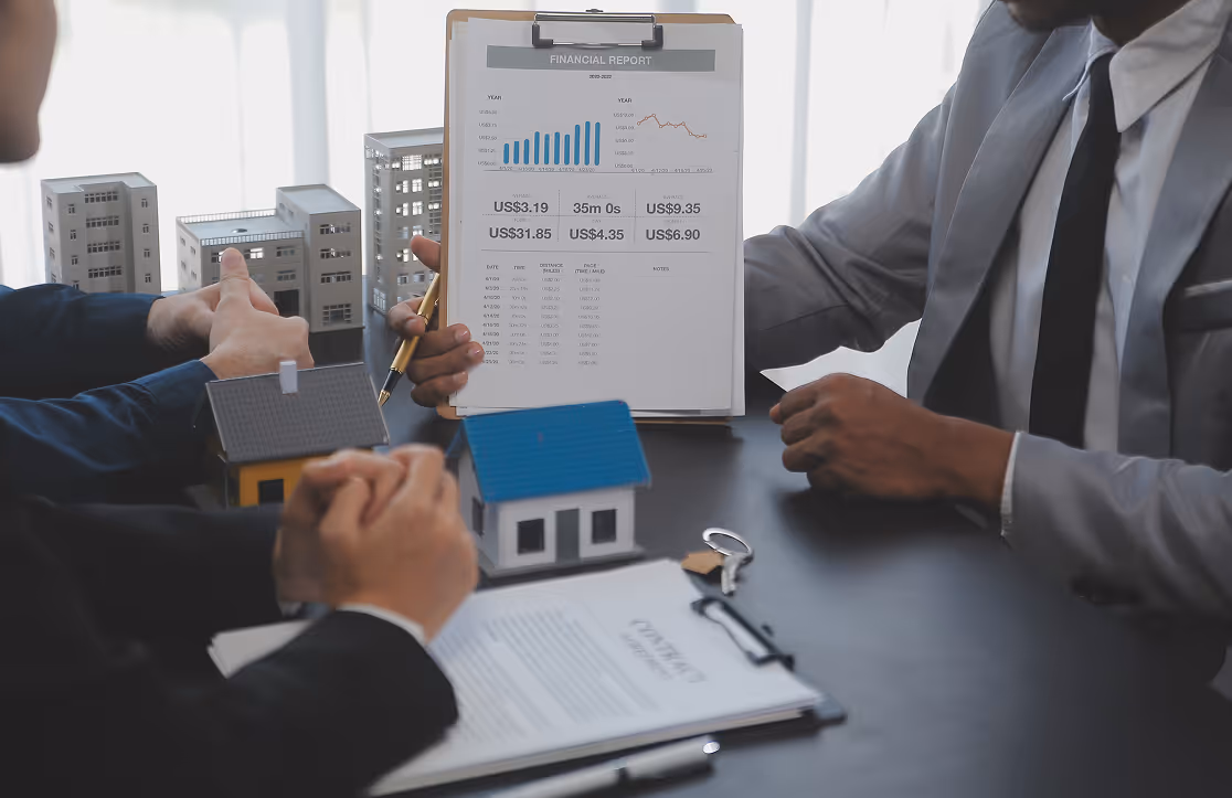 Two men review a financial report with charts beside real estate models and a contract on a table.