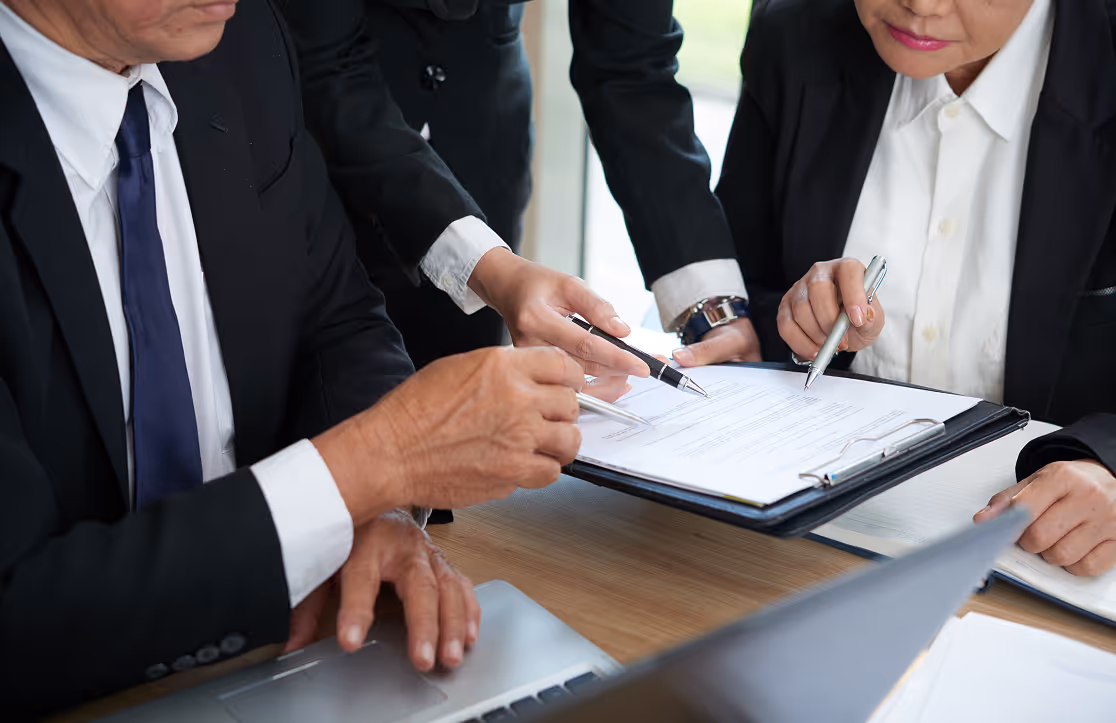Three business professionals in suits reviewing and pointing at documents on a clipboard over a wooden desk.