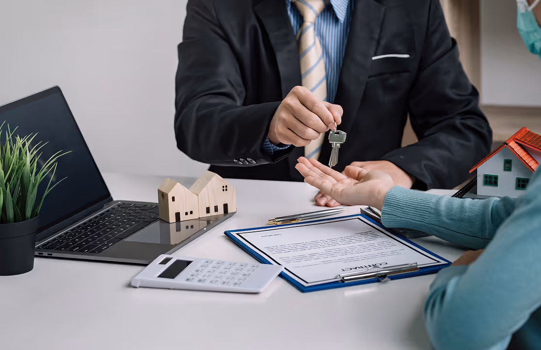 A person in a suit handing over house keys to another person at a desk with contract papers and models.