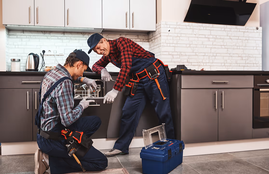 Two repairmen in uniforms fixing a dishwasher in a modern kitchen with tools and toolbox nearby.