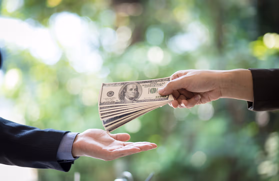 Hand passing several hundred-dollar bills to another open hand outdoors with blurred green background.