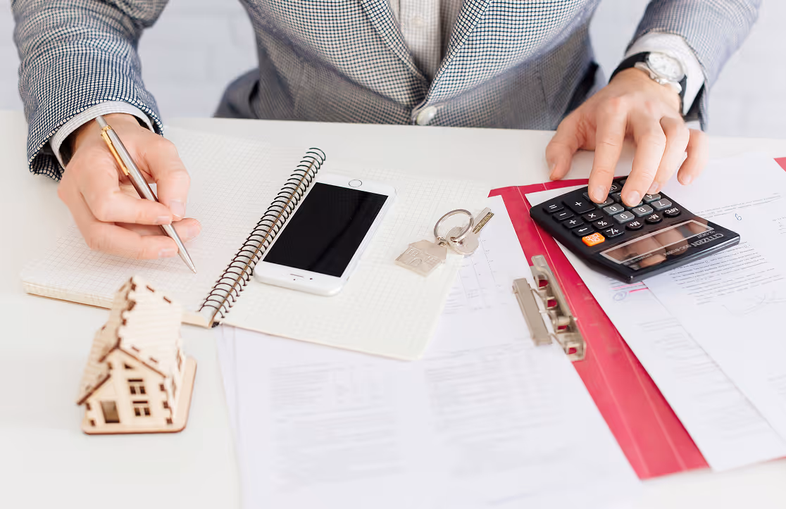 Person using calculator and writing notes with house model, smartphone, and keys on table.