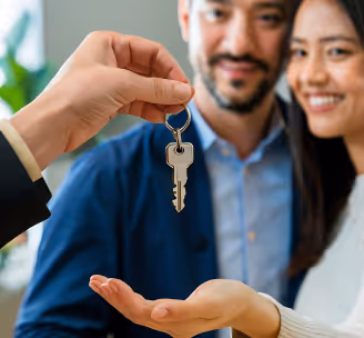 Hand giving a key to a smiling couple receiving it with open hands indoors.