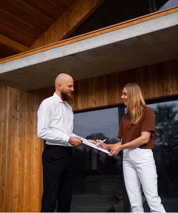 Man and woman smiling while holding clipboard outside a wooden modern building.