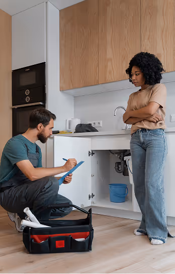 Technician notes maintenance details while inspecting kitchen cabinet plumbing with concerned woman watching.