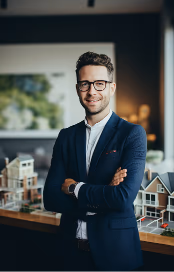 Smiling man in glasses and blue suit stands with arms crossed near property model houses indoors.