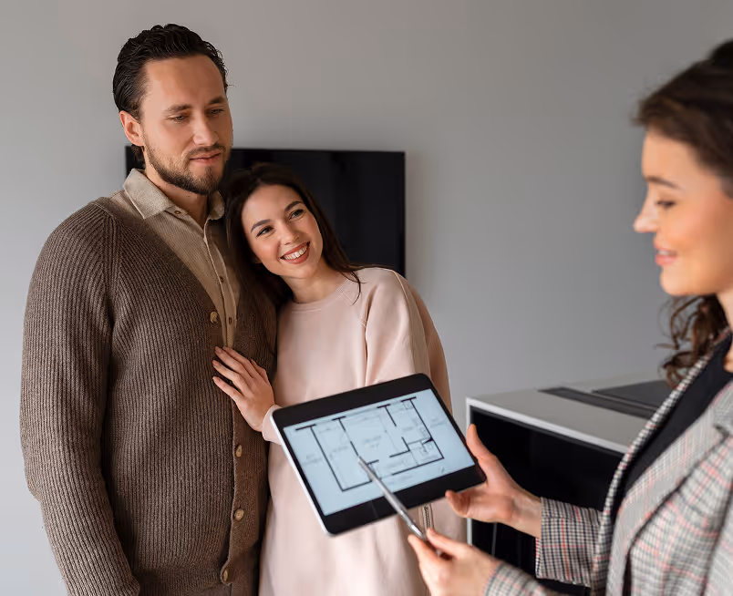 Real estate agent shows floor plan on tablet to couple standing close together indoors.