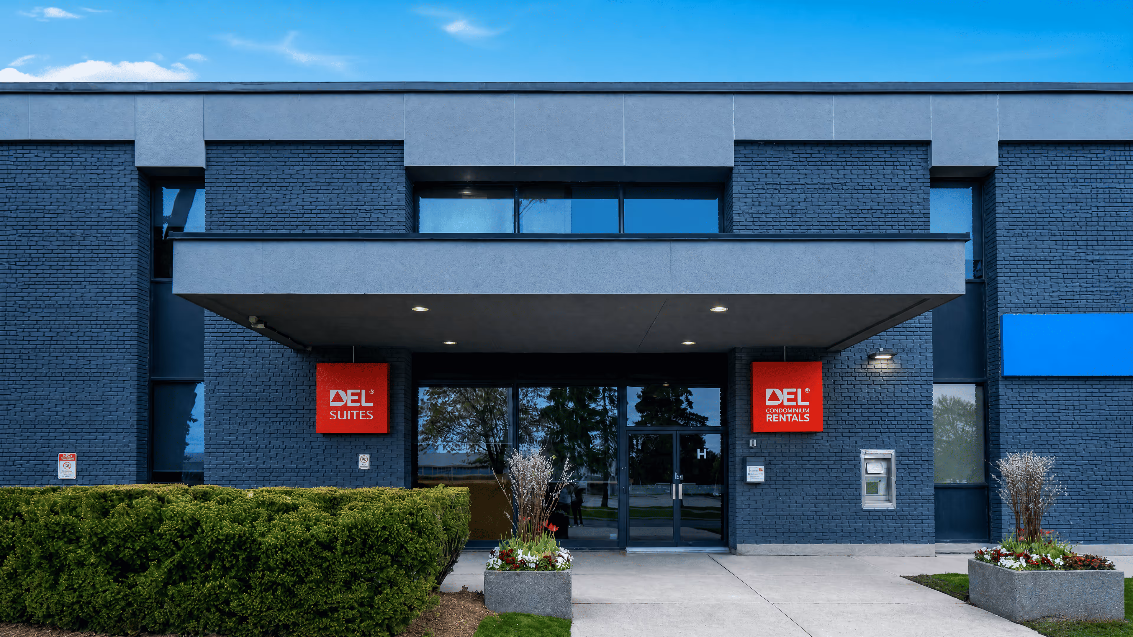 Entrance of a modern building with dark brick walls and two red signs reading DEL Suites and DEL Rentals.