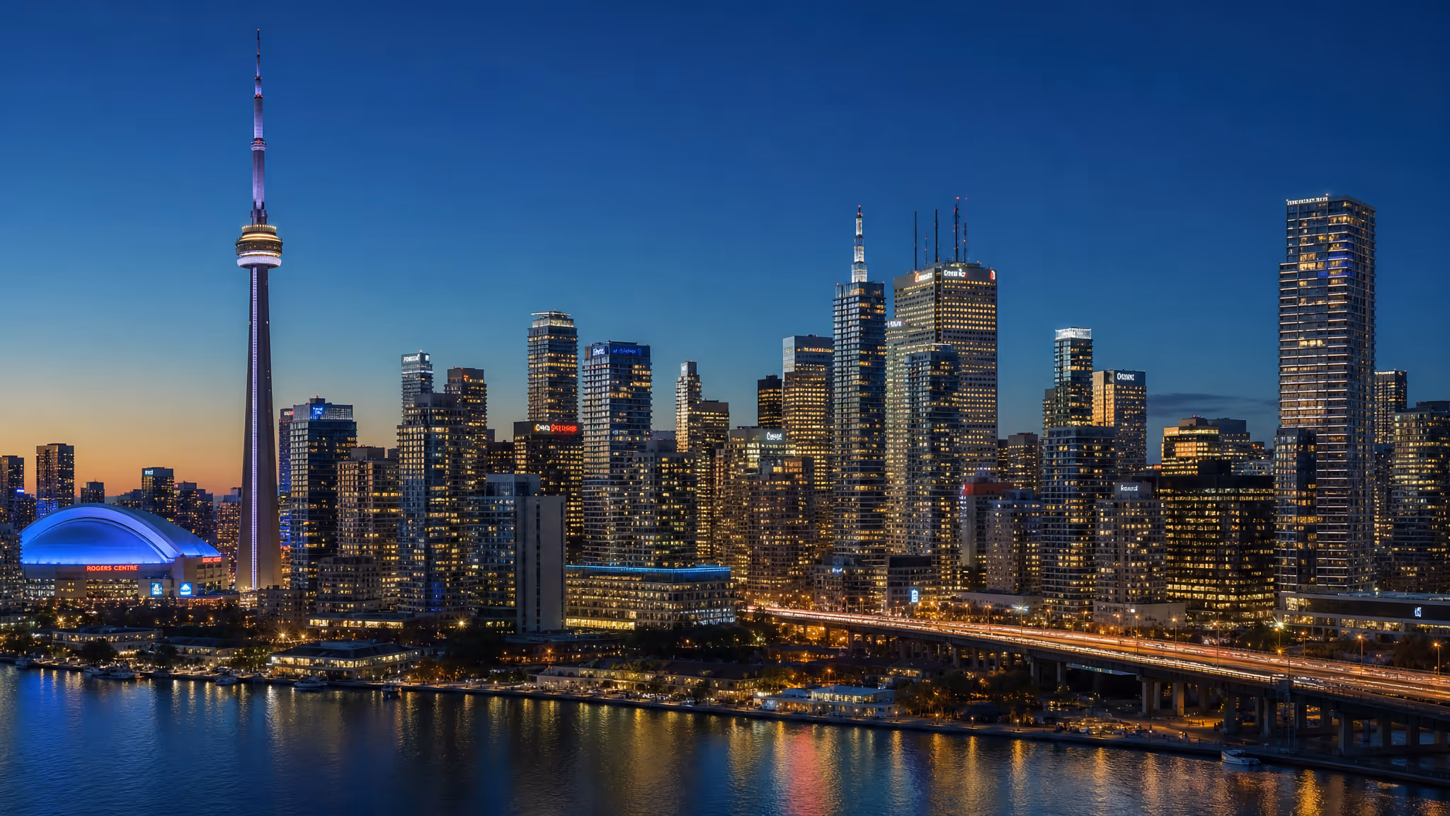 Toronto skyline at dusk with CN Tower, Rogers Centre, lit skyscrapers, and waterfront reflecting lights.