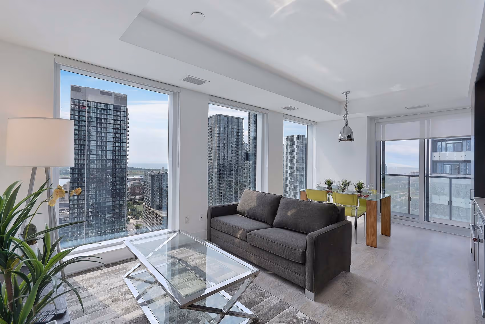 Modern apartment living room with gray sofa, glass coffee table, dining area, and tall windows with city views.