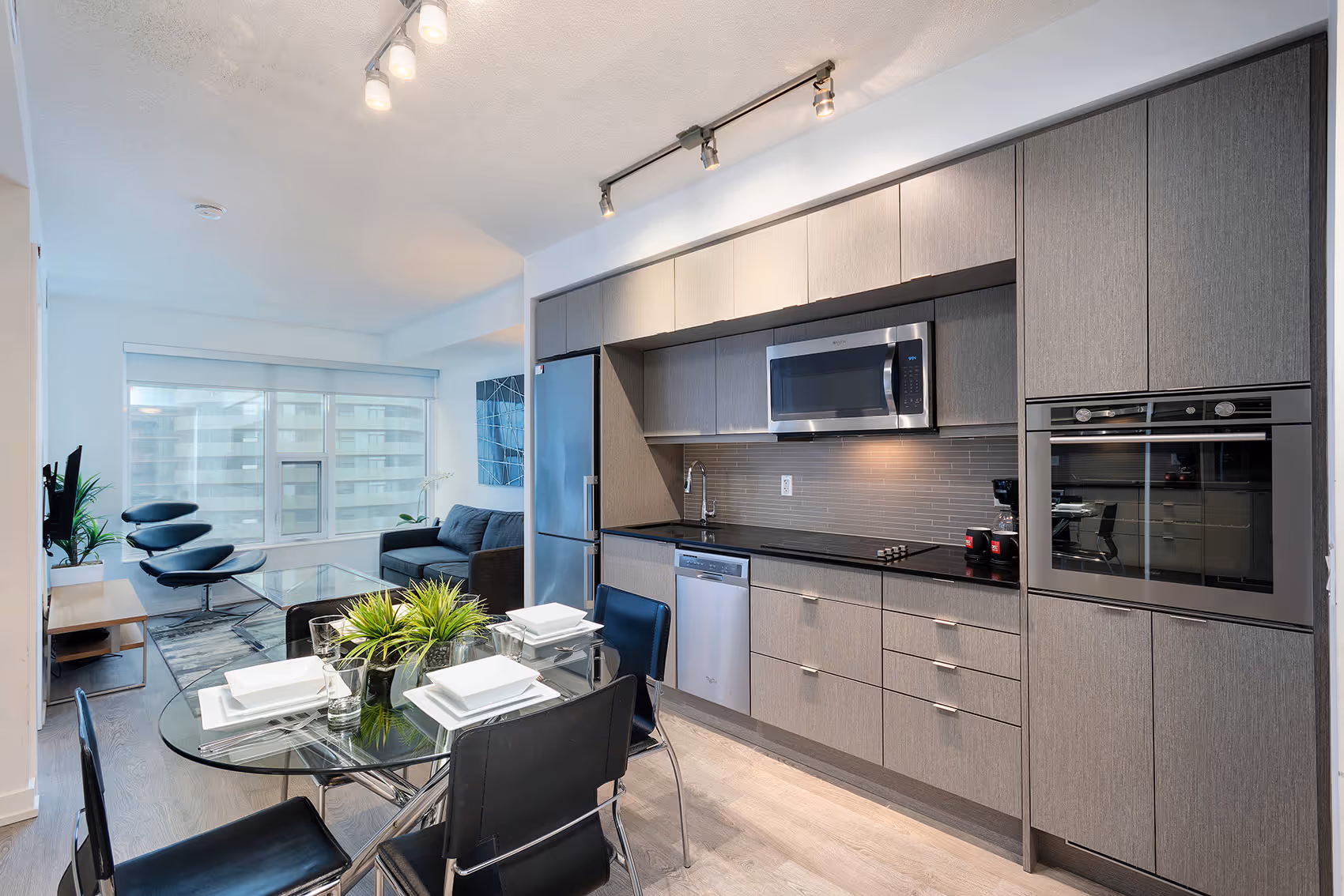 Modern kitchen and dining area with glass table, black chairs, gray cabinets, and a living room seating area.