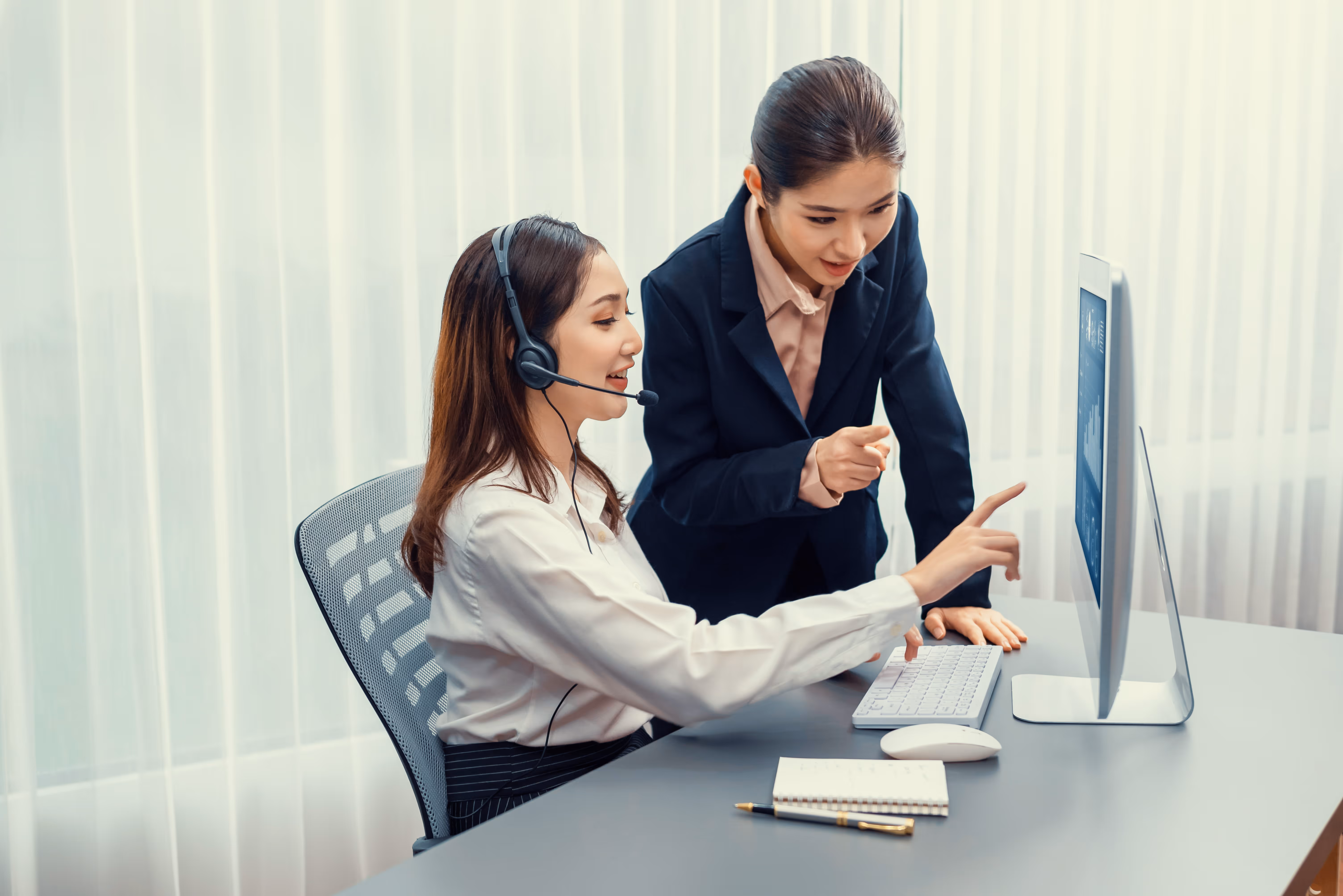 Two women looking at a computer screen, one wearing a headset and sitting, the other standing and pointing.