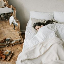 Woman sleeping in a white bed with a large ornate mirror and a tray with tea and snacks on a wooden floor nearby.