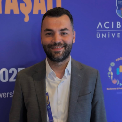 Smiling man with short dark hair and beard wearing a gray suit and white shirt, standing in front of a blue background with text and logos.