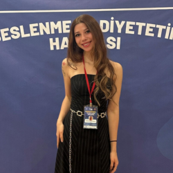 A young woman with long wavy hair wearing a black strapless dress and a red lanyard standing in front of a blue background with partially visible white text.