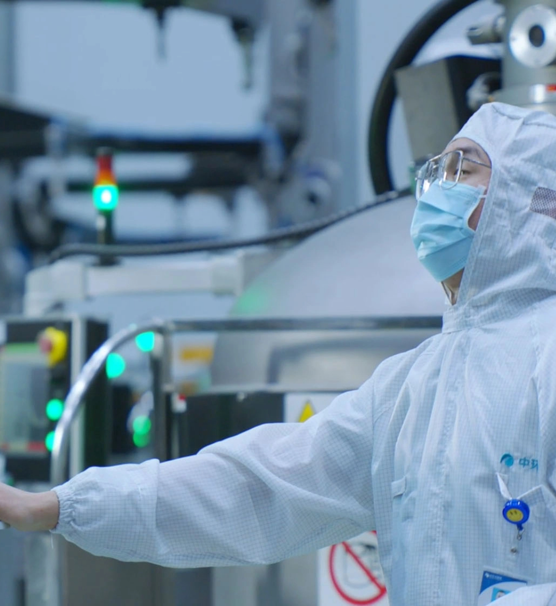 Worker in a cleanroom suit, mask, and safety glasses operating machinery in a high-tech manufacturing environment.