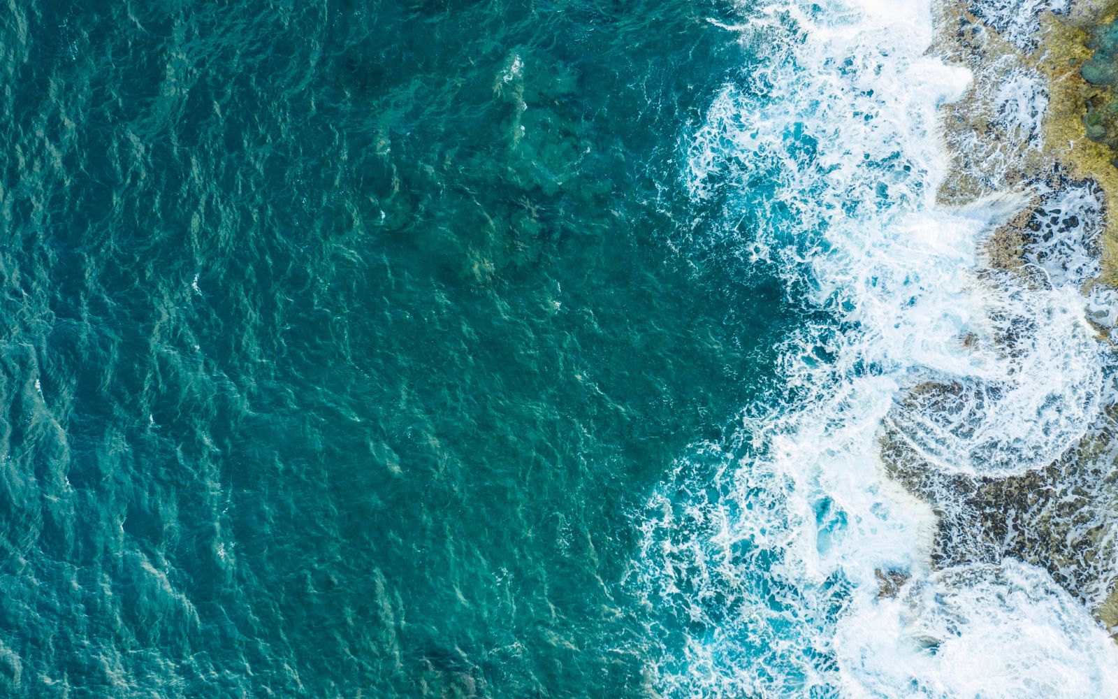 Aerial view of ocean waves crashing onto rocky shore with white foam and turquoise water.