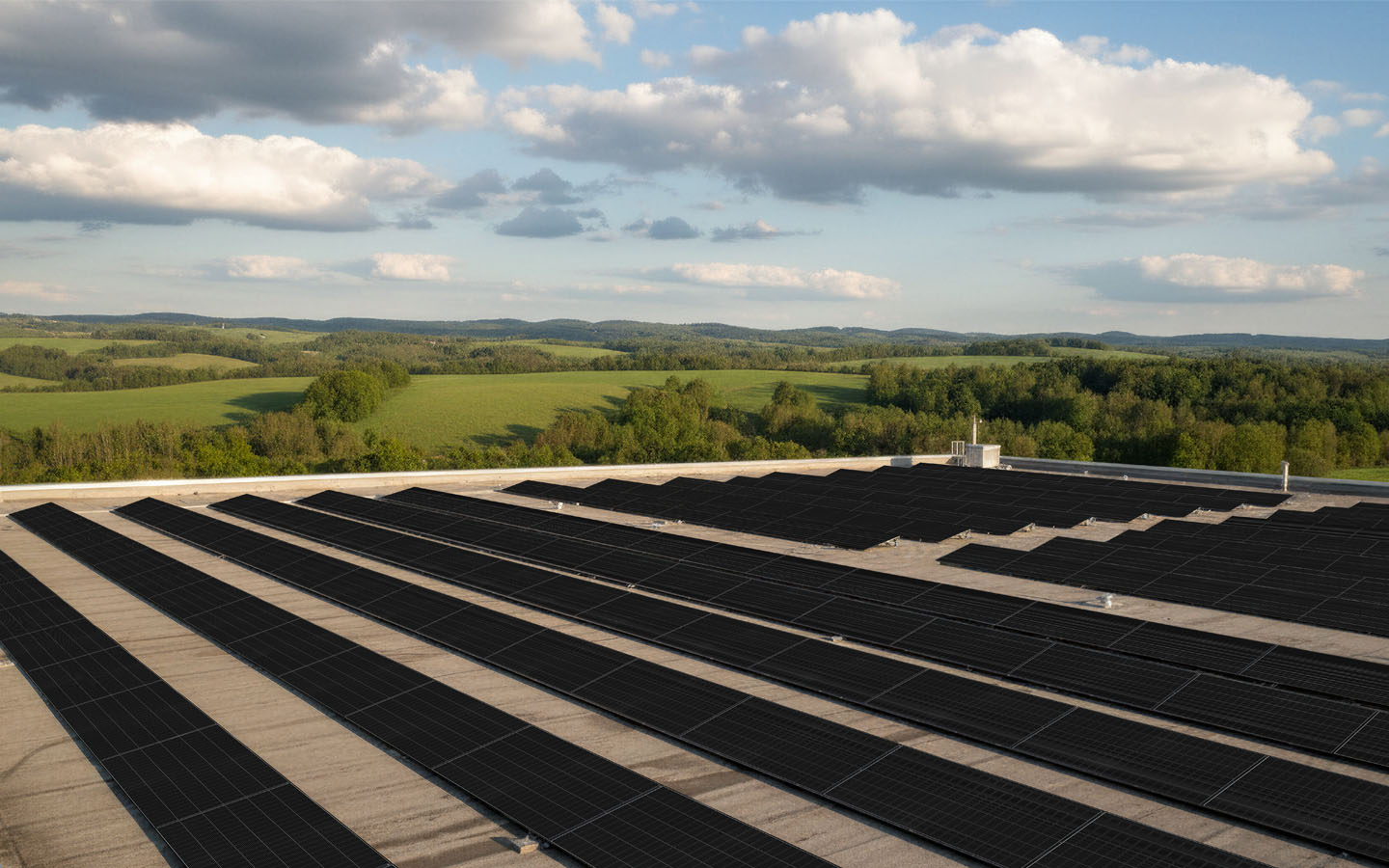 Rows of black solar panels installed on a flat roof with green hills and a partly cloudy sky in the background.