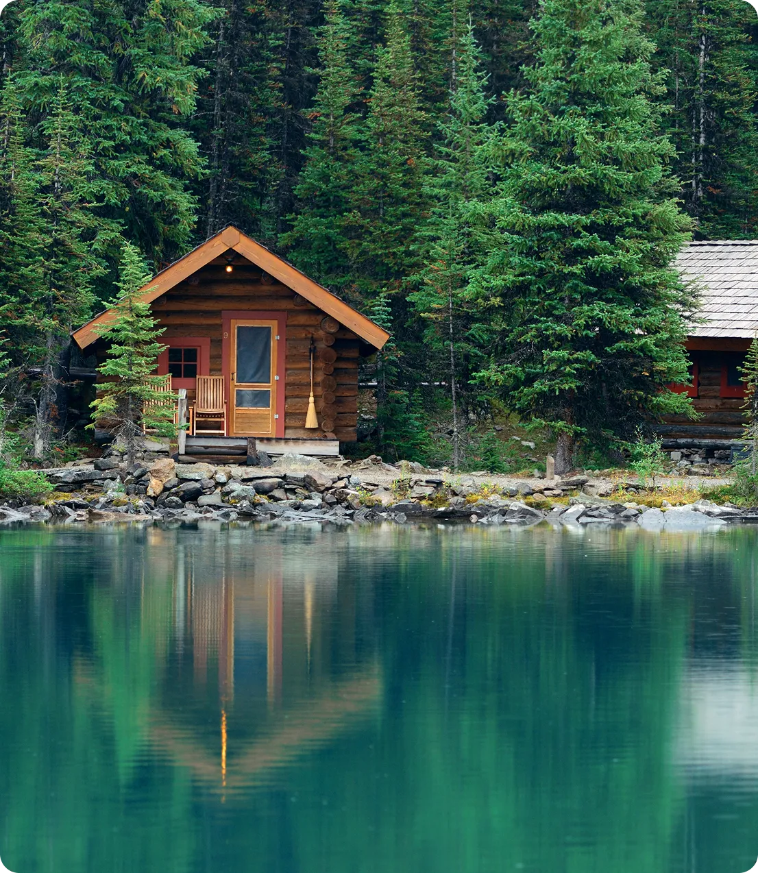 Small wooden cabin with rocking chair by a calm lake surrounded by dense evergreen trees.