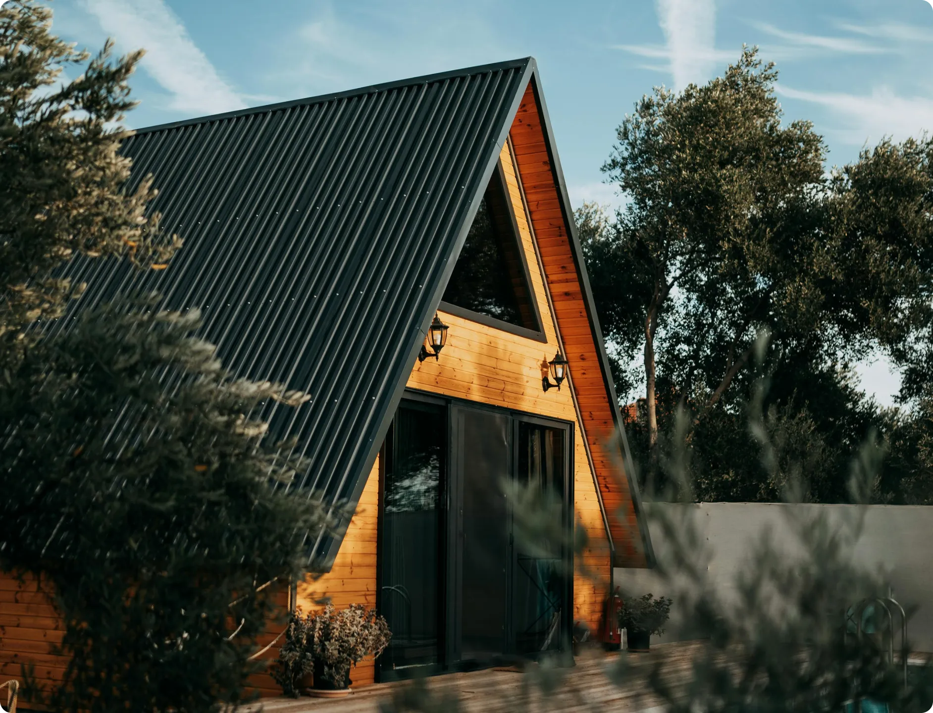 Wooden A-frame cabin with black metal roof surrounded by trees and plants in natural light.