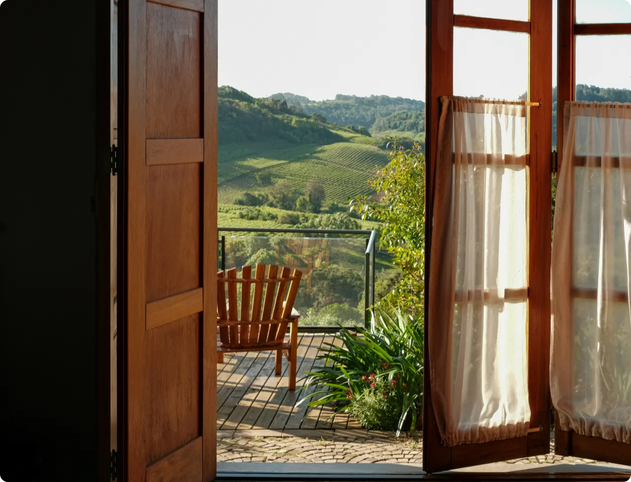 Open wooden door and glass door with sheer curtains leading to a balcony with a wooden chair overlooking green hills.