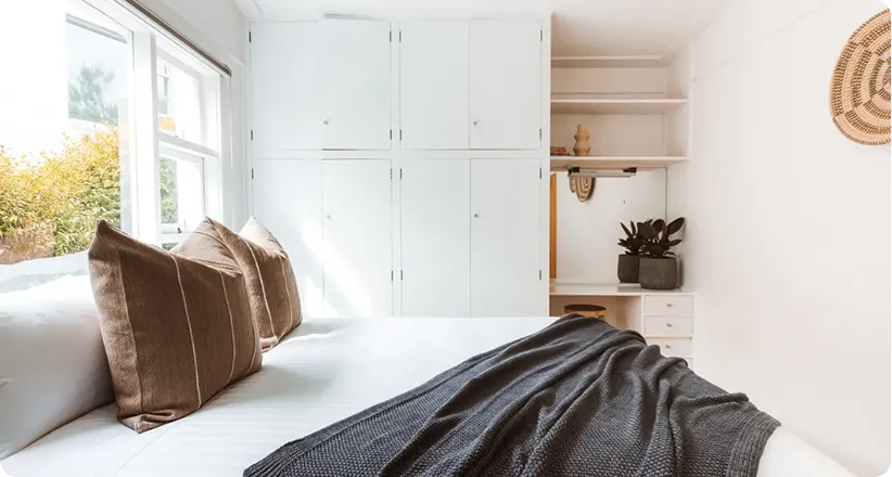 Bright bedroom with white walls, large window, white bed with brown pillows, gray blanket, and white cabinets with shelves.