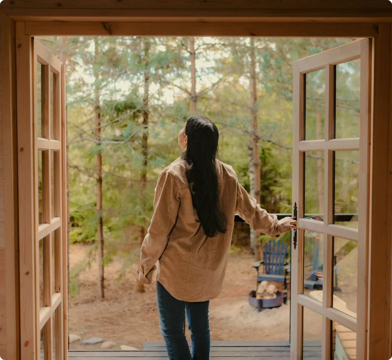 Person with long dark hair standing on a wooden porch, opening double glass doors to look at a forest landscape.