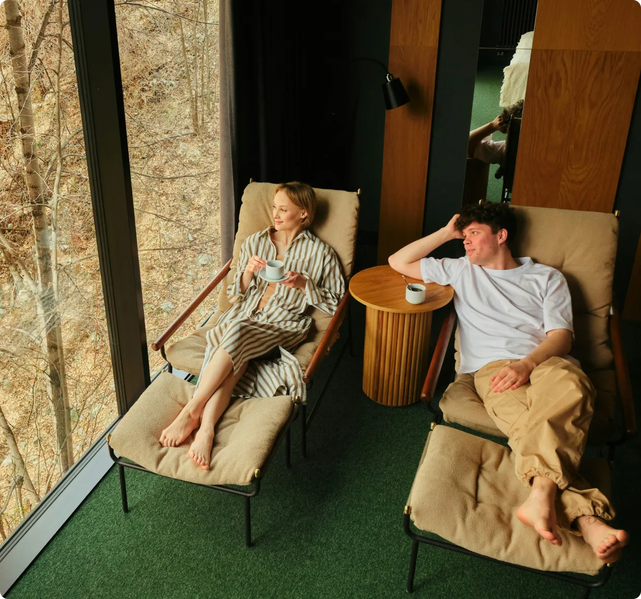 A woman and a man relaxing barefoot on cushioned lounge chairs indoors near a large window overlooking bare trees, with the woman holding a cup and the man resting his arm on a round wooden side table with a cup.