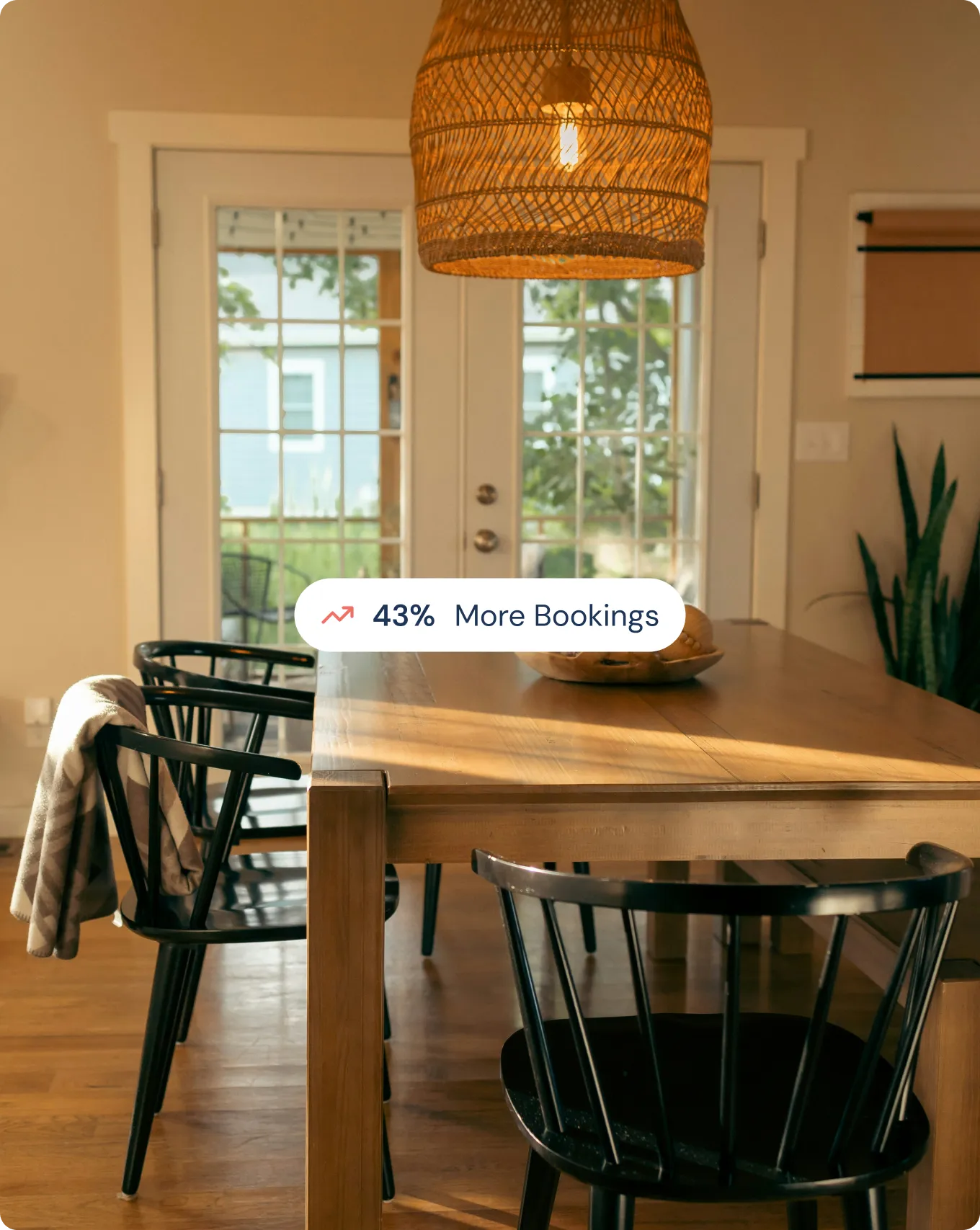 Dining room with a wooden table, black chairs, a woven pendant light, and sunlight coming through glass doors.