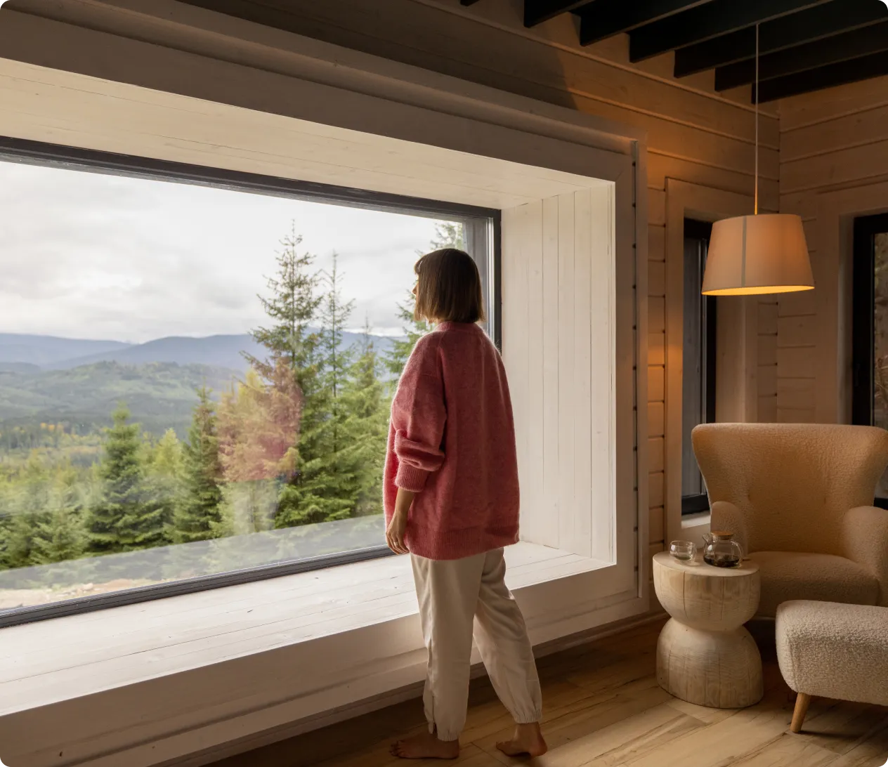 Woman in a pink sweater standing barefoot by a large window, looking out at a forested mountain landscape.