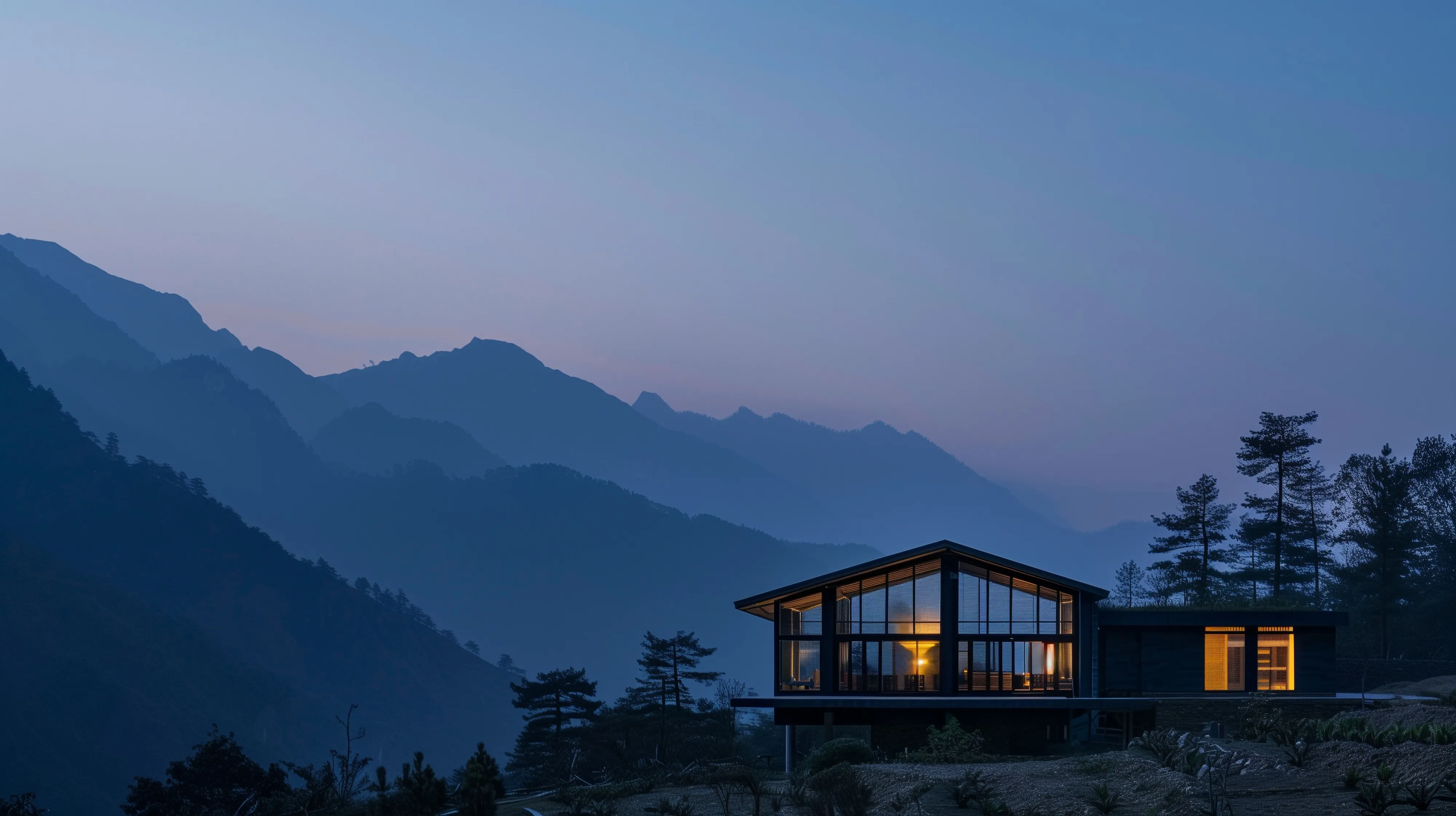 Modern house illuminated at dusk set against layers of blue mountain silhouettes and trees.