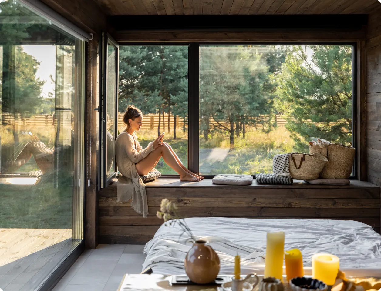 Woman sitting on a wooden window seat in a cozy room, looking at her phone with a forest view outside.