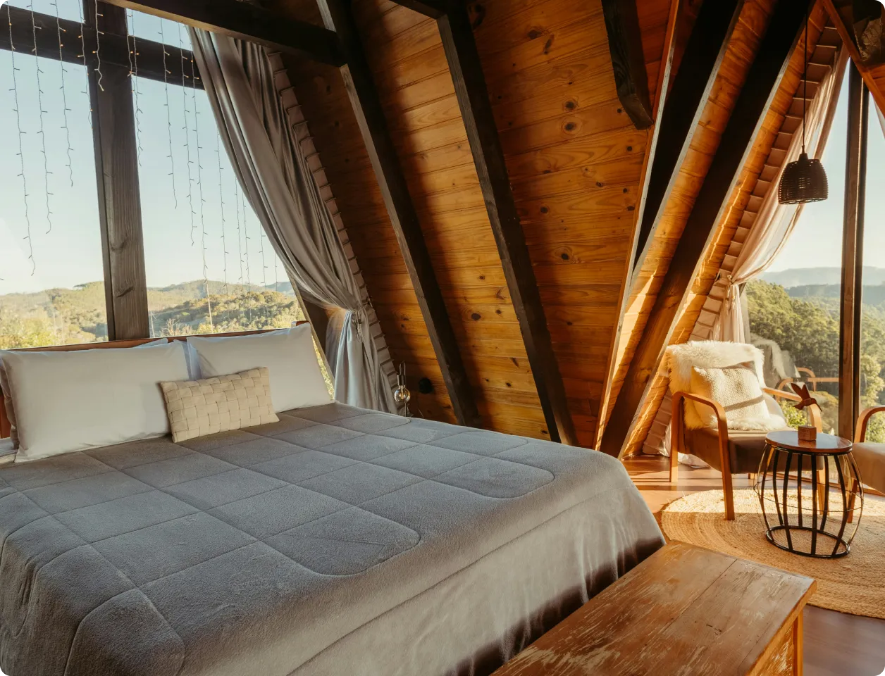 Cozy wooden A-frame bedroom with large bed, gray bedding, armchair with fur throw, round table, and windows showing a forested mountain view.